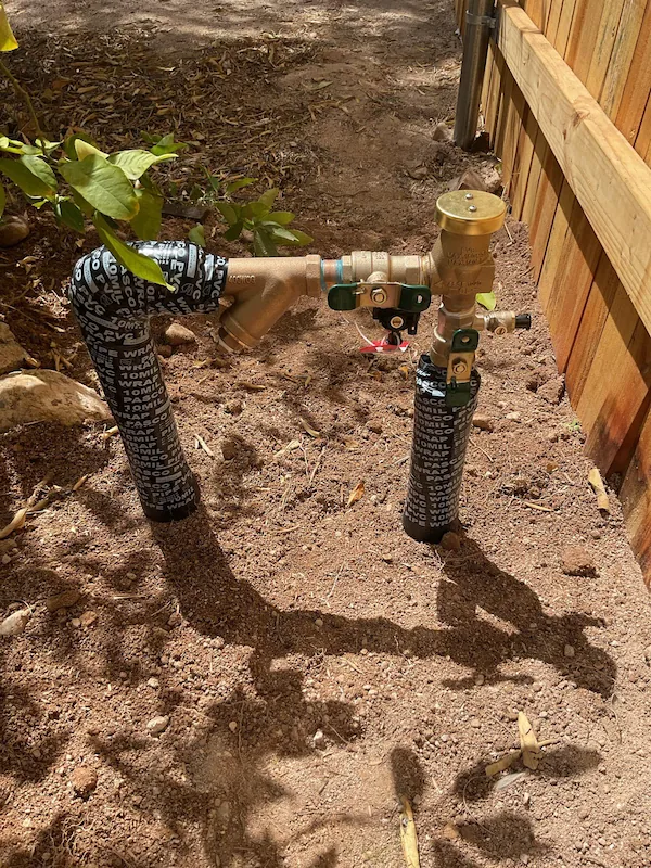 Outdoor brass commercial plumbing pipes with valves and insulation next to wooden fence in dirt ground.