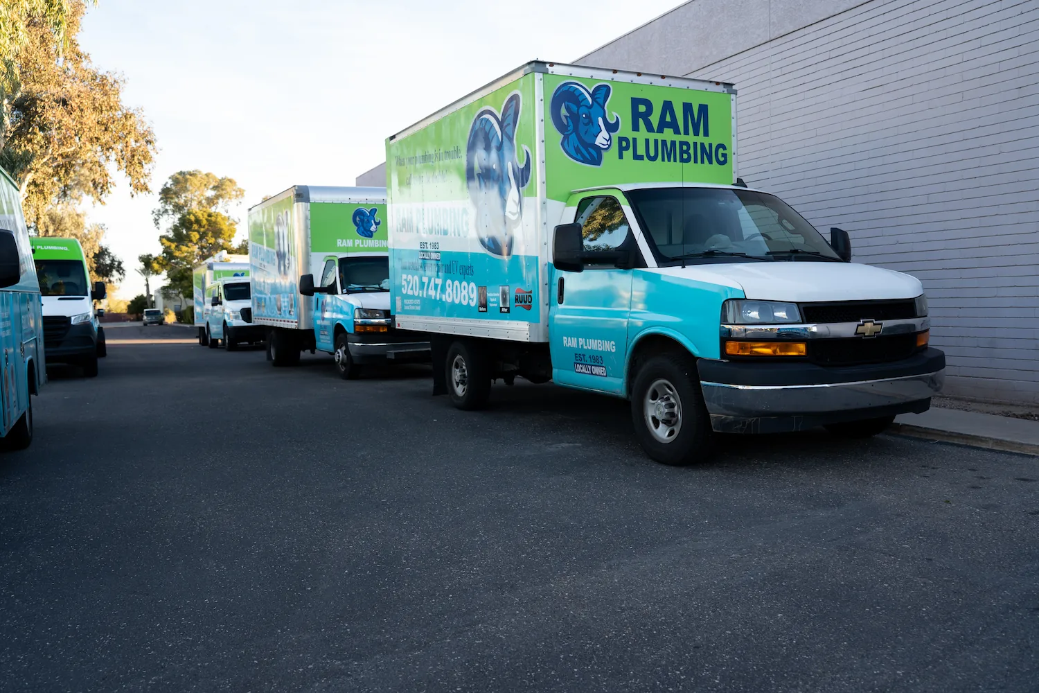 Line of Ram Plumbing service trucks parked beside a building on a street at dusk.