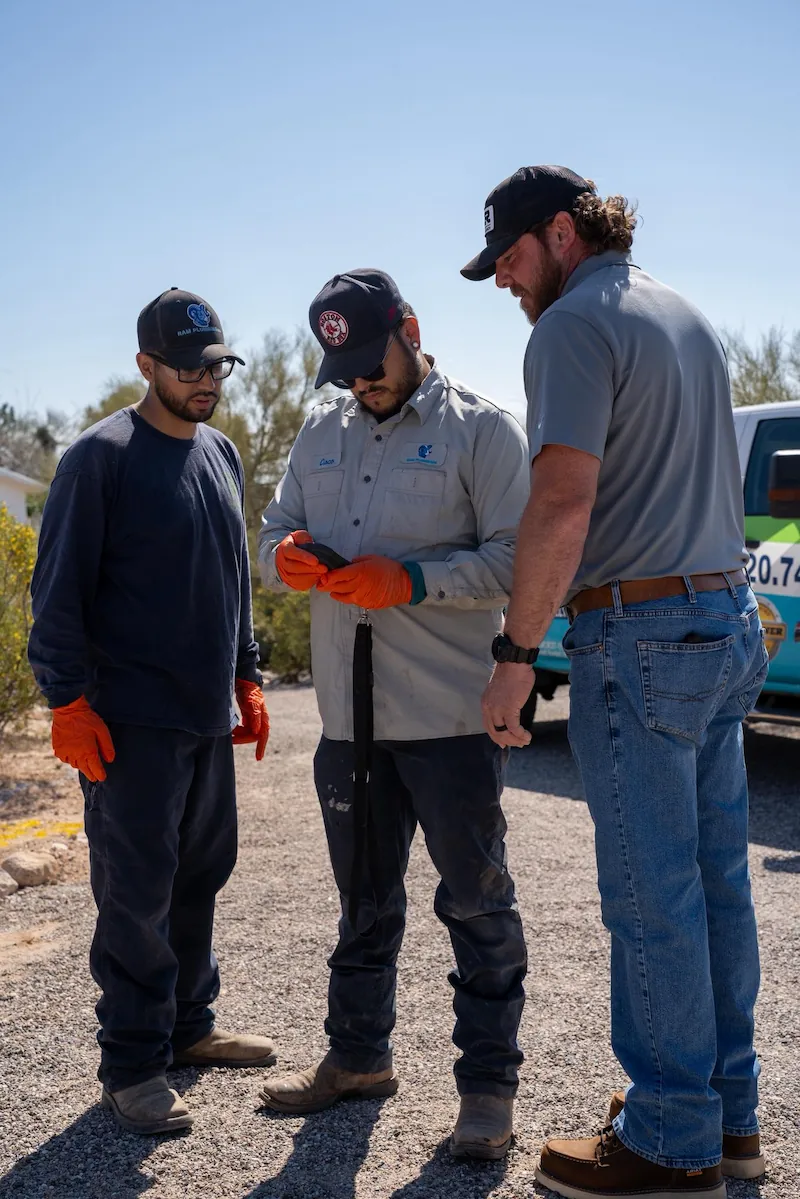 Three workers wearing caps and gloves standing outside on gravel looking at a phone.