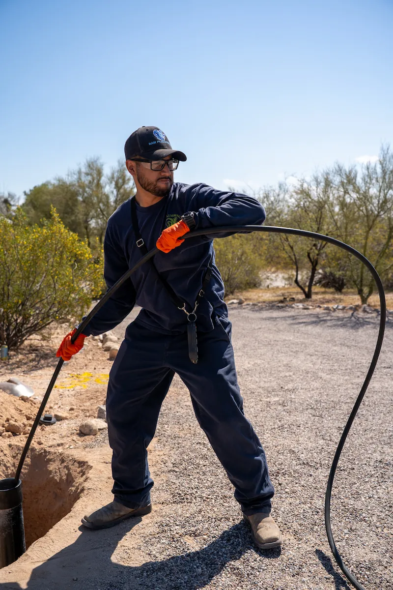 Man in a cap and gloves handling a long black hose by a trench on a gravelly area with trees.