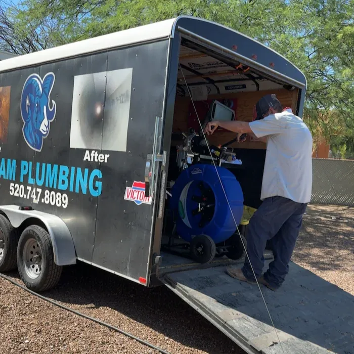 Man working with equipment inside a plumbing service trailer parked outdoors on gravel.