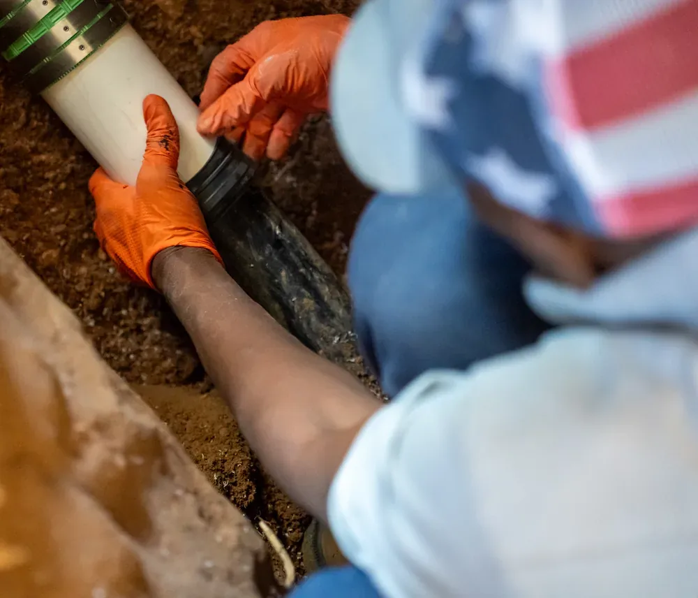 Person wearing orange gloves installing a white pipe in soil, seen from overhead.