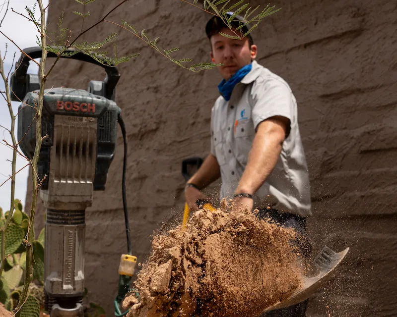 Man shoveling dirt near a Bosch jackhammer against a brick wall outdoors.