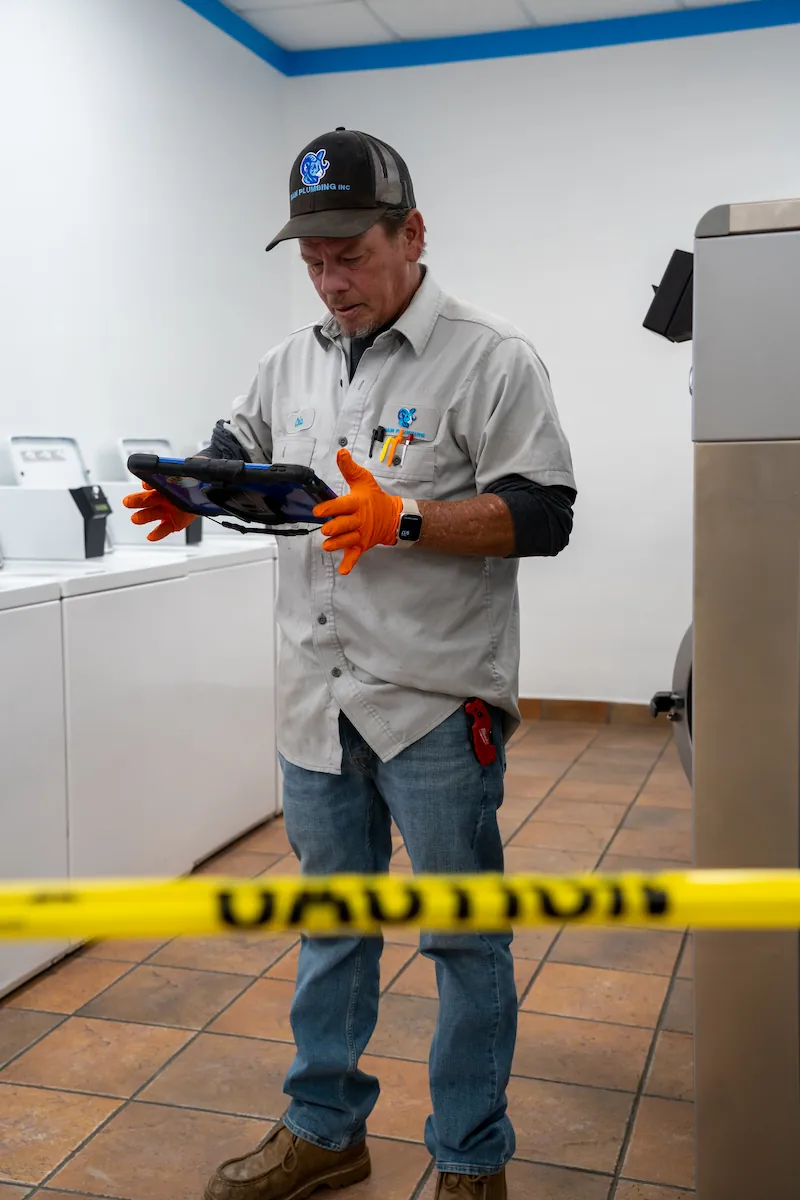 Plumber wearing gloves and uniform reads tablet behind caution tape in laundry room with machines.