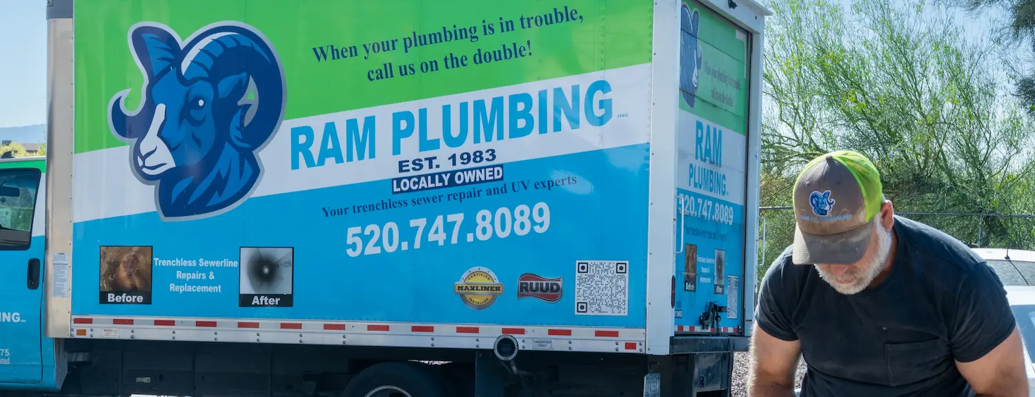 Man in RAM Plumbing cap working near a RAM Plumbing service truck with contact info and logo.