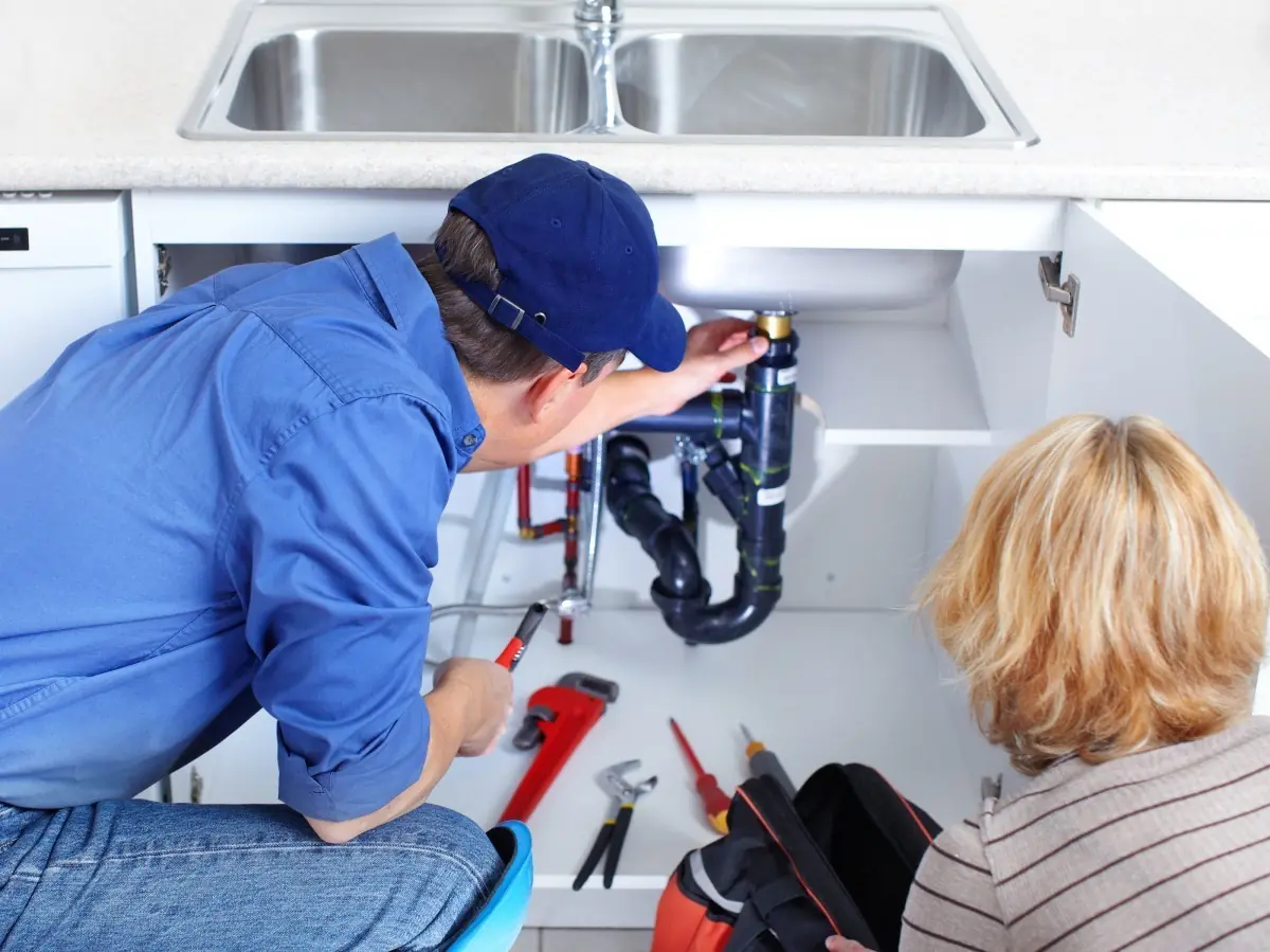Plumber repairing kitchen sink pipes under cabinet while woman watches.