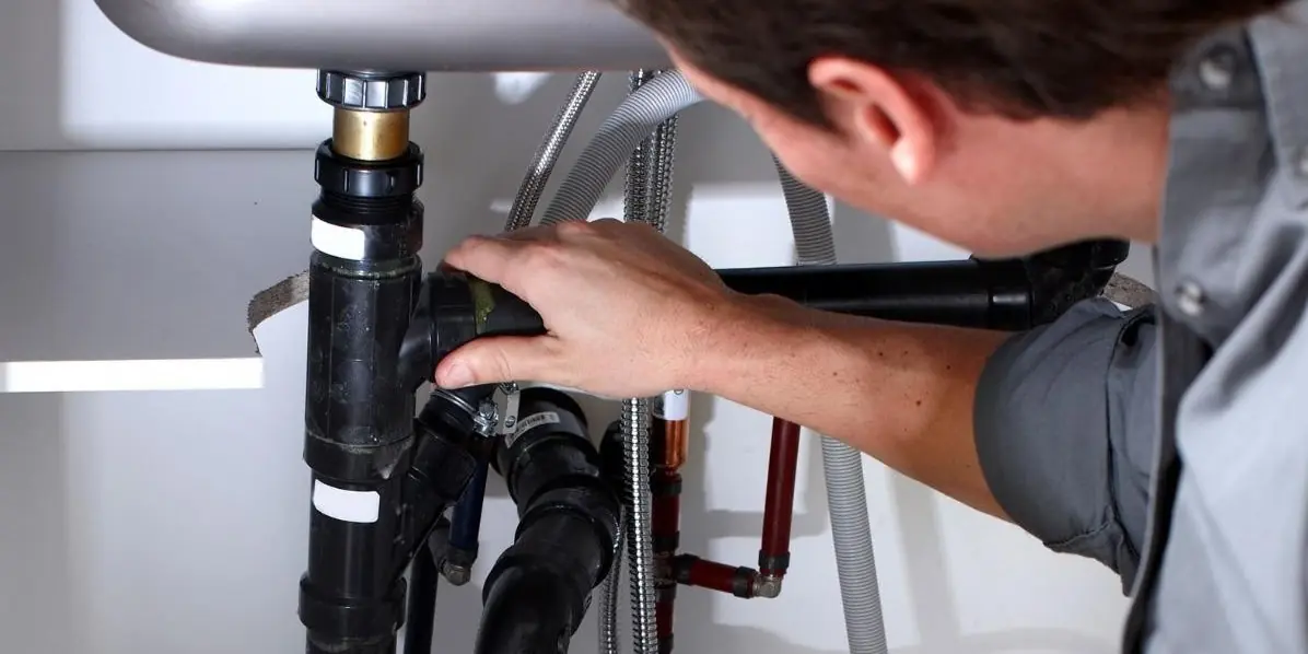 Plumber inspecting and tightening black pipes under a sink with water supply lines visible.