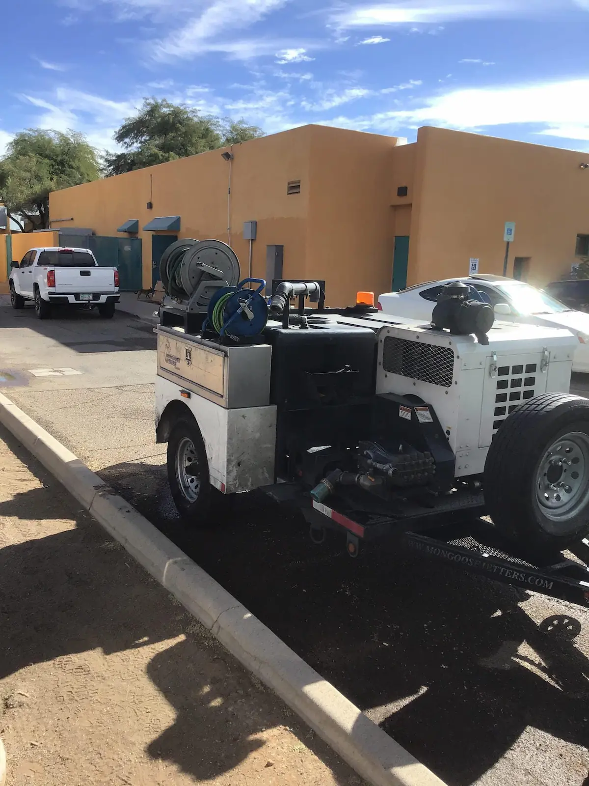 Pressure washer equipment trailer parked on street near beige building under blue sky.