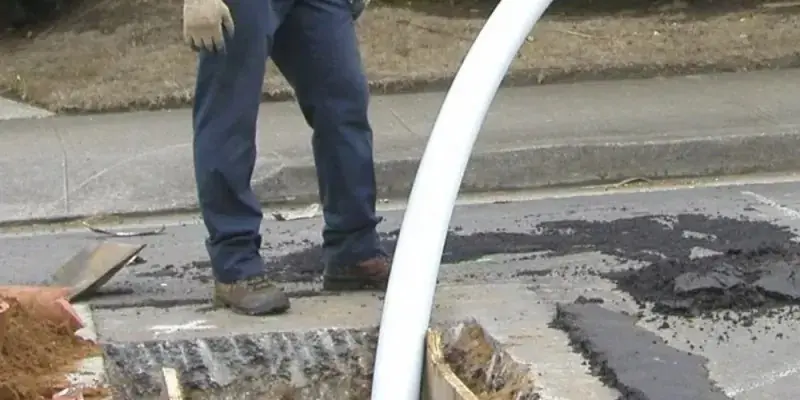 Worker in blue pants and gloves standing near a dug-up street with construction materials.