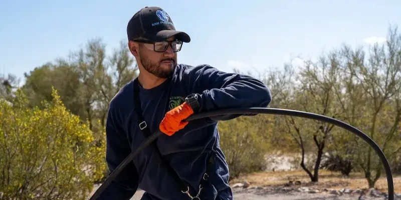 Man wearing glasses and gloves handling a large black hose outdoors with trees and blue sky.