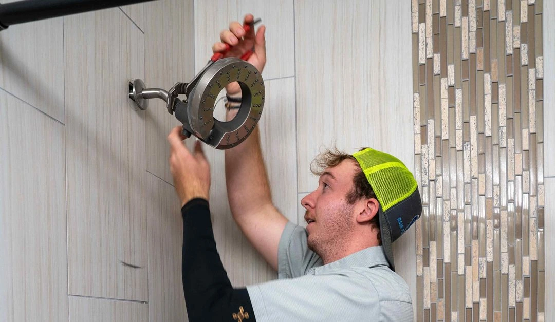 Worker wearing cap installing or adjusting a circular shower head on a tiled bathroom wall.