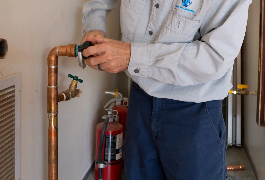 A plumber in a gray shirt working on a copper pipe with a fire extinguisher in the background.