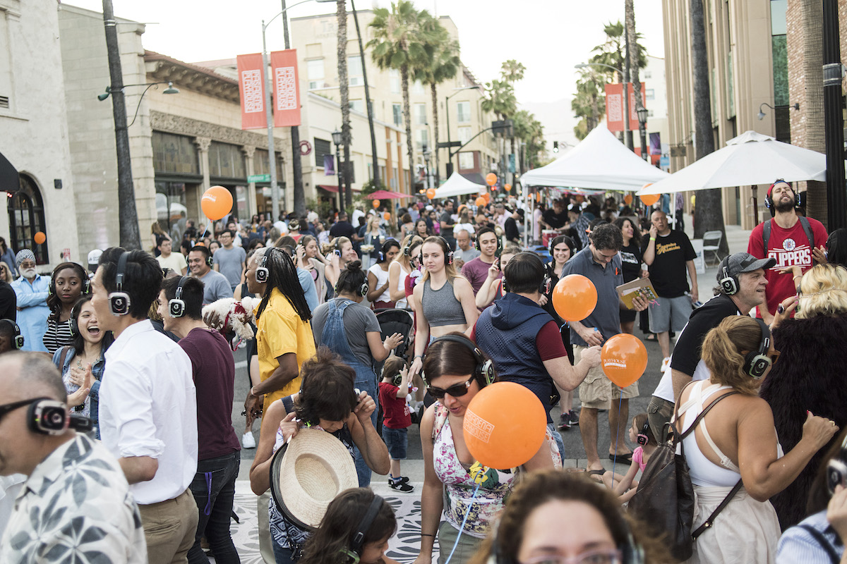 A large, diverse crowd fills a blocked-off street at an outdoor event. Many attendees wear wireless headphones. Orange balloons and white event tents line the street, with palm trees and commercial buildings visible in the background.