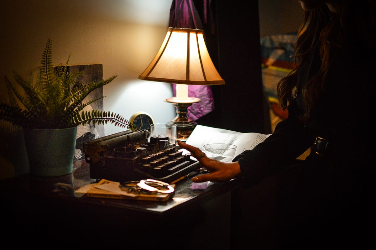 A dimly lit desk scene with a vintage black typewriter at center. A person's hand rests on a sheet of paper beside the typewriter. A glowing table lamp illuminates the surface, which also holds a small clock, handcuffs, a glass, and a potted fern.