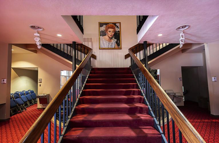 The lobby of a theater, viewed from the base of a carpeted central staircase. A large portrait of a woman in a white dress hangs on the wall at the top of the stairs. Red carpet extends across the floor, and rows of blue seats are visible.