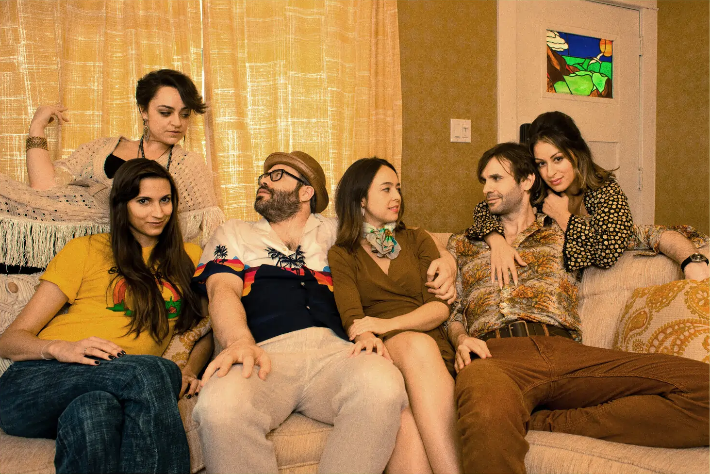 Six people in 1970s-style clothing lounge on and around a couch in a warmly lit retro living room. The group poses casually, some seated and some standing, against a backdrop of gold curtains, patterned wallpaper, and a stained glass window panel.