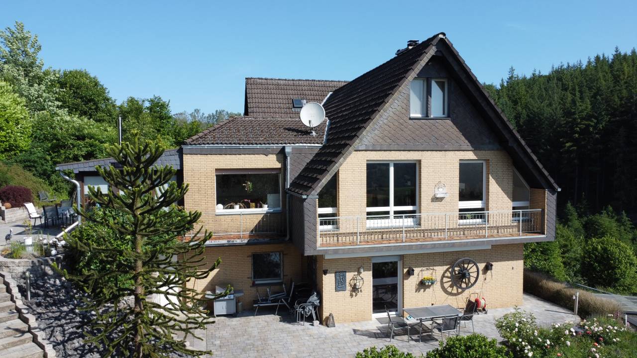 Großes zweistöckiges Haus mit Balkon, Terrasse, Garten und umliegenden Bäumen unter klarem blauem Himmel.