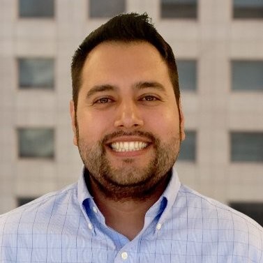 Smiling man with dark hair and beard wearing light blue button-up shirt indoors