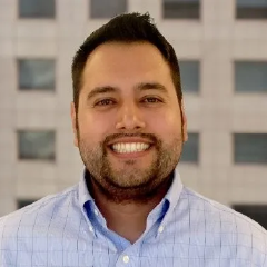Professional headshot of smiling man in light blue striped shirt outdoors
