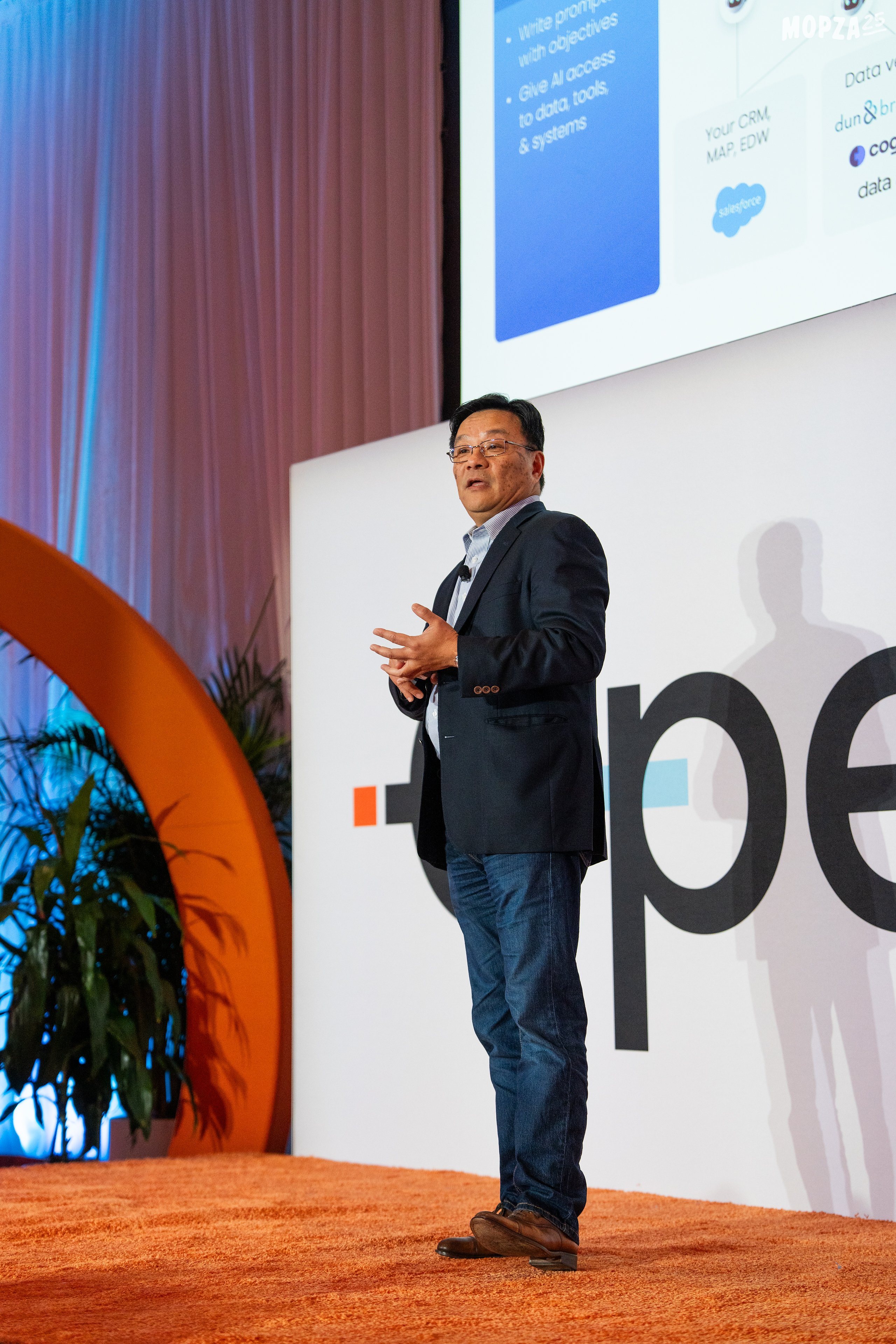 Man in business casual attire presenting on stage with projection screen and large text display