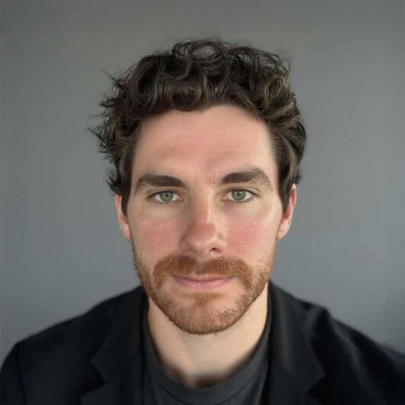 Portrait of a man with dark wavy hair, beard, and green eyes against gray background.