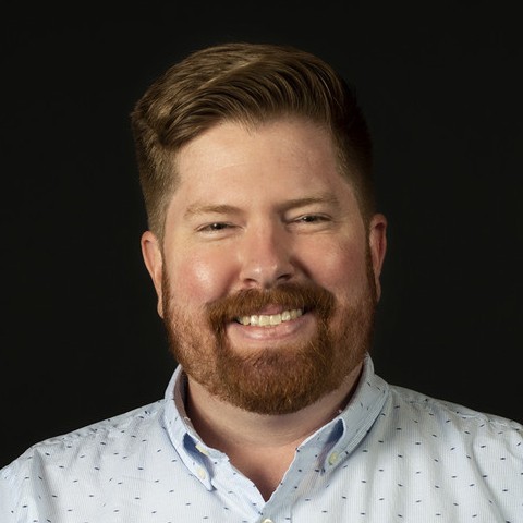 Smiling man with brown beard wearing light blue patterned shirt against black background