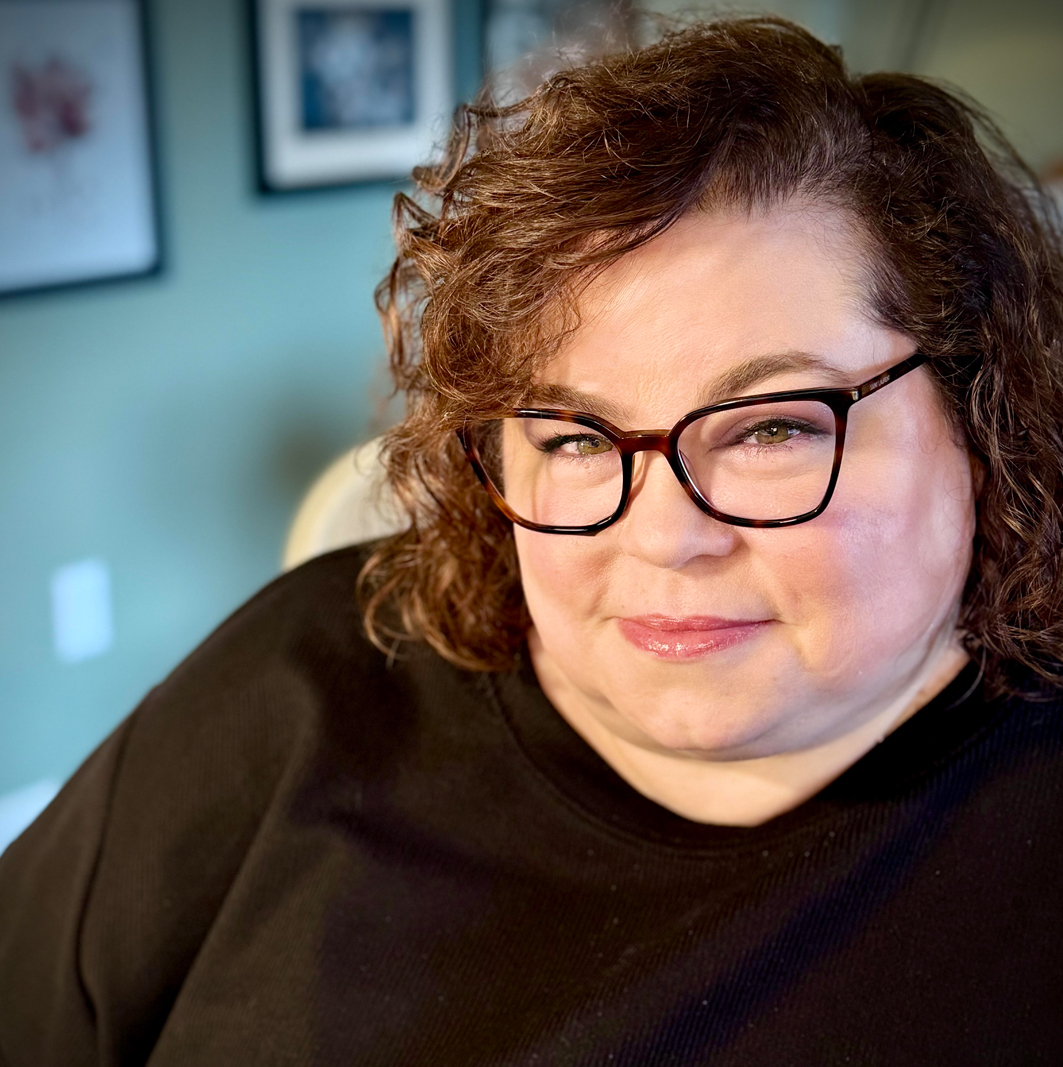 Portrait of person with curly reddish-brown hair wearing black glasses and black shirt.