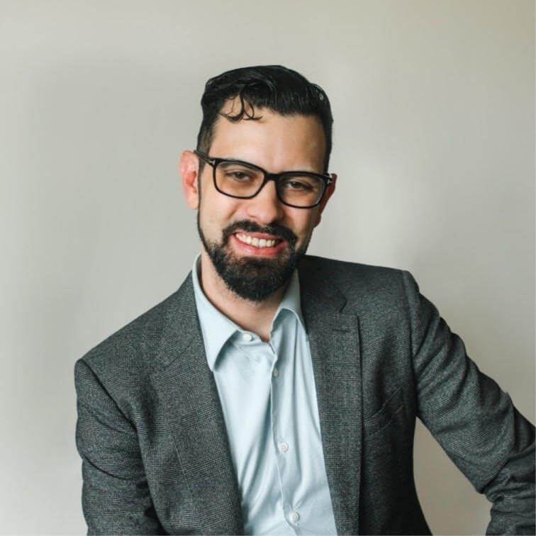Bearded man wearing glasses and dark blazer smiling at camera