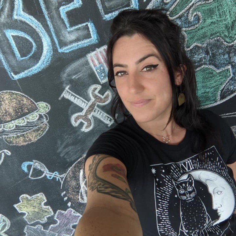 Woman with dark hair and arm tattoo posing against colorful chalk drawings.