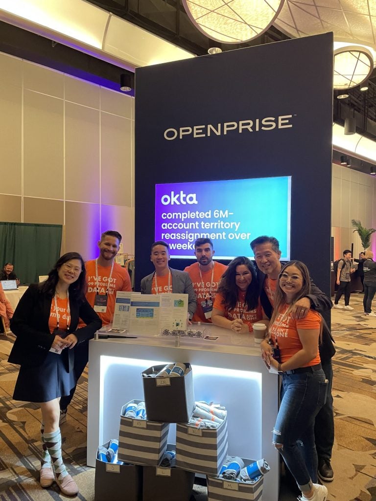 OpenPrise and Okta team members at conference booth with branded signage and merchandise display