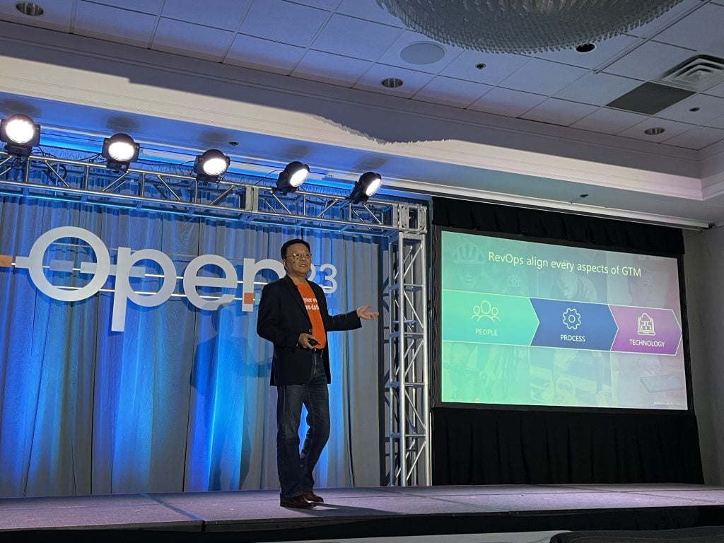 Speaker presenting on stage about DevOps with screen showing process and technology frameworks.