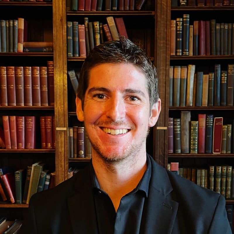 Smiling man in dark blue shirt posed before wooden bookshelf filled with books