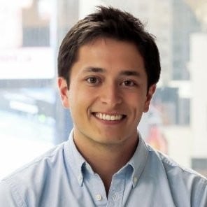 Smiling man in light blue shirt with city skyline visible through window behind