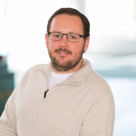 Man wearing glasses and beige sweater smiling at camera indoors