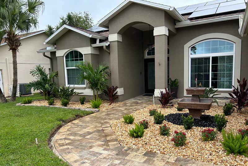 front yard of a modern house with a curved brick walkway, landscaped with small shrubs, palm trees, and a stone water fountain in tampa bay