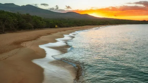 Sunset over a long empty beach, kiawe trees and a dark volcanic ridge rising inland