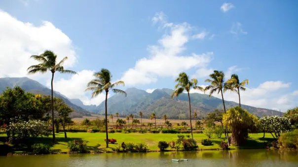 Palms and flowering gardens beside a still pond, West Maui Mountains ridged behind under blue sky