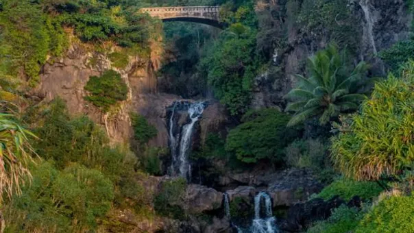 Stacked waterfalls drop through a green gorge beneath an arched highway bridge, ridges rising behind