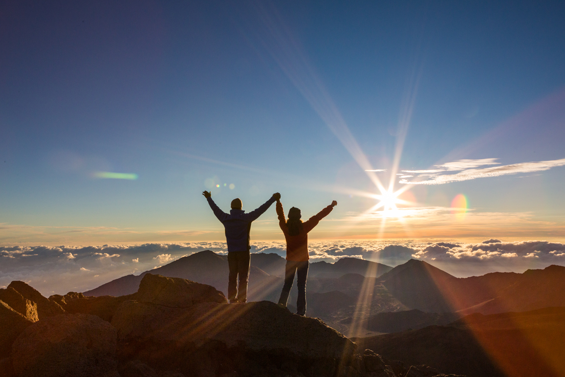 Two people with arms raised at sunrise on the Haleakala summit above the clouds