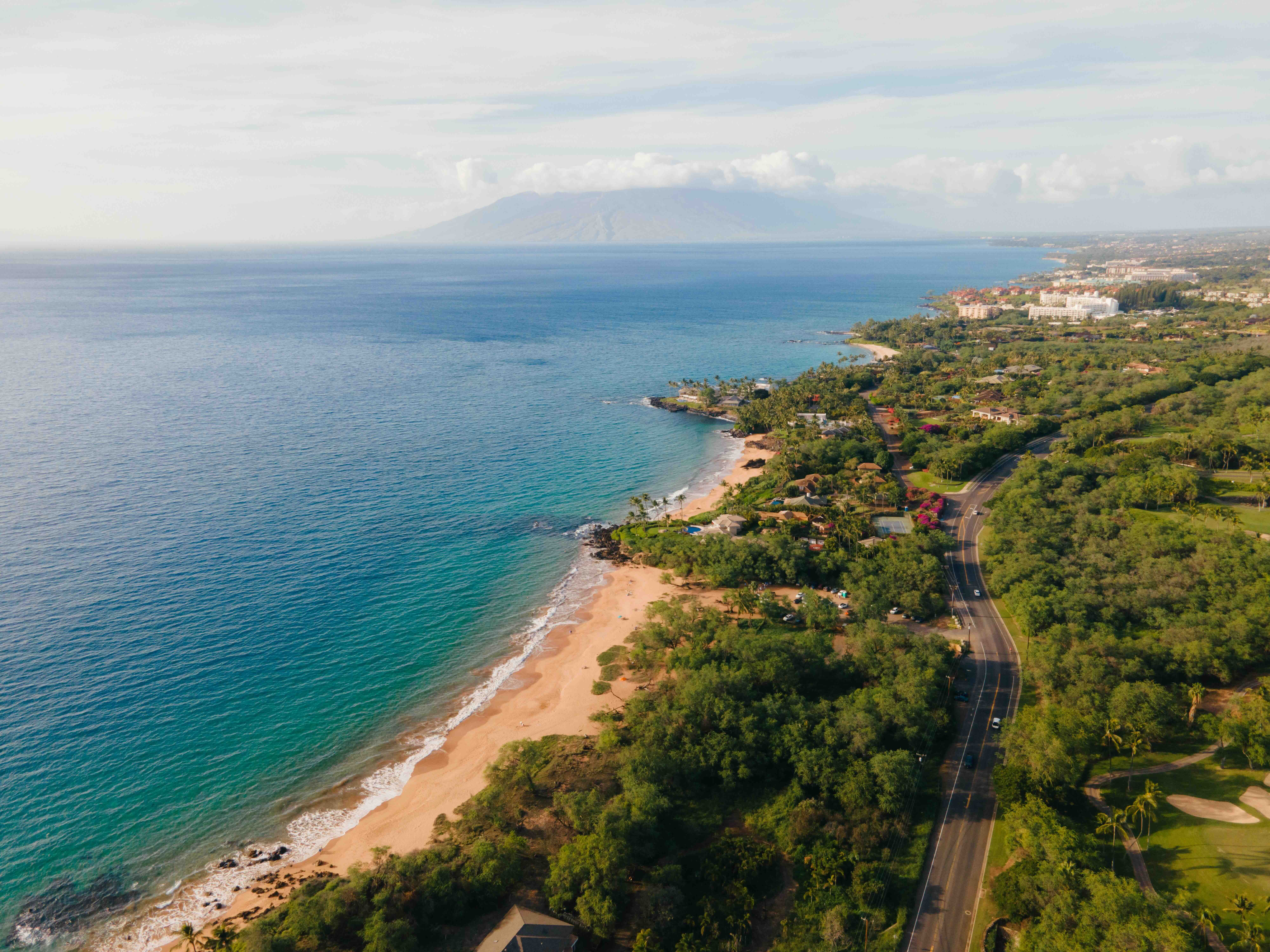 Aerial view of a South Maui road running between green hillside and a sandy cove
