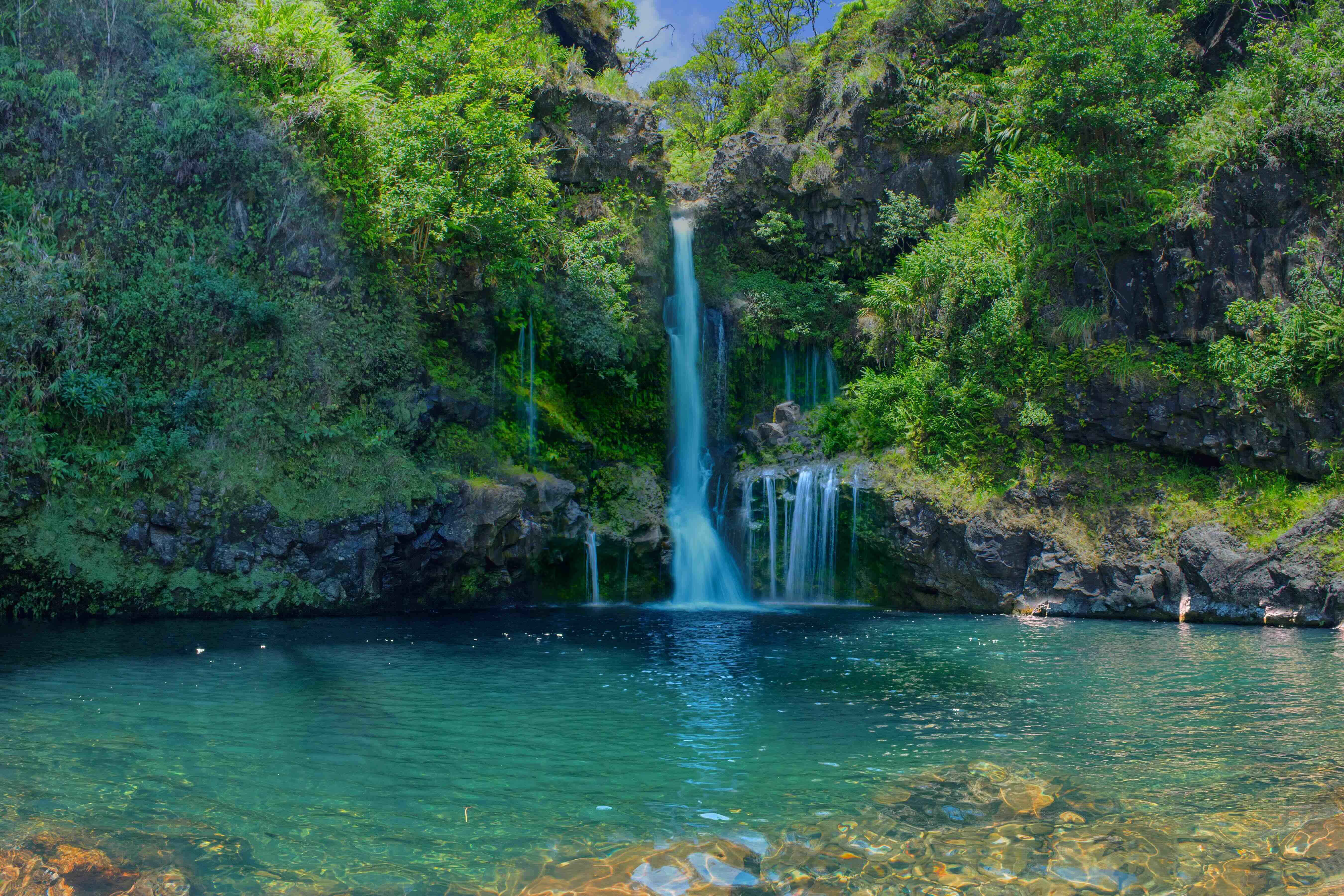 A tiered waterfall dropping down a mossy rock face into a clear turquoise pool ringed by dense green forest