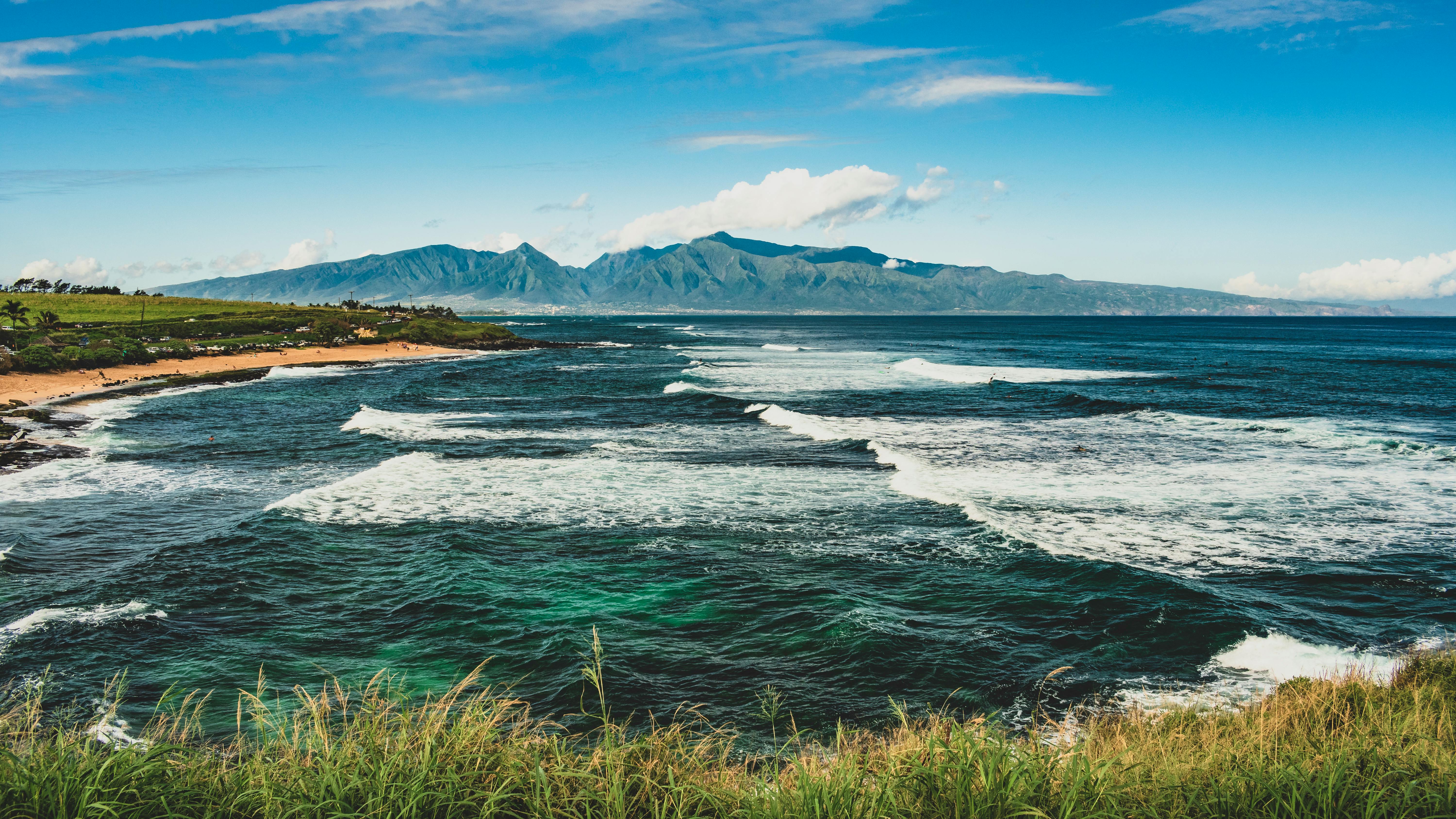 Surf breaking in long white lines across Hoʻokipa Bay on Maui's north shore, with grass headland in the foreground and the West Maui Mountains beyond