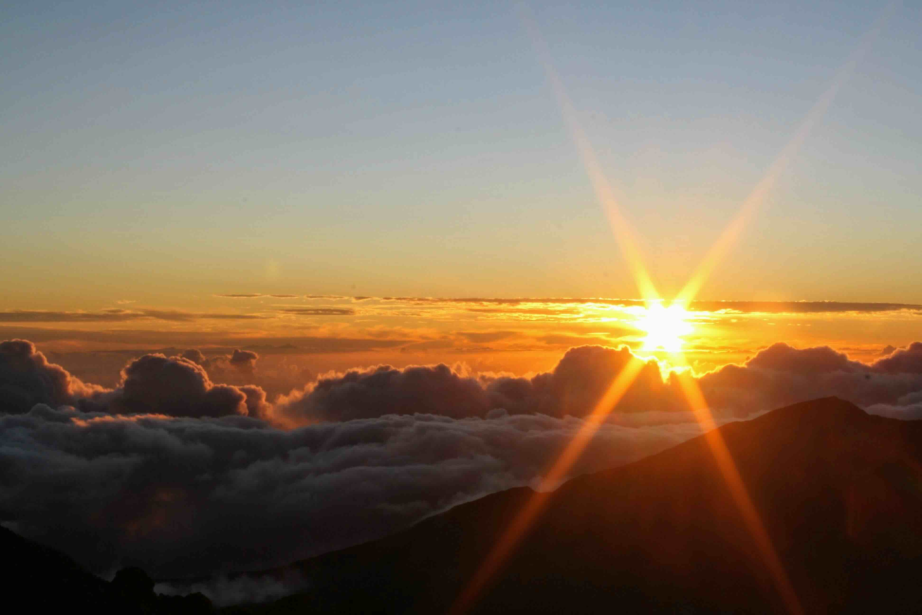 The sun low on the horizon above a deck of cloud, seen from the Haleakalā summit with the crater rim in silhouette