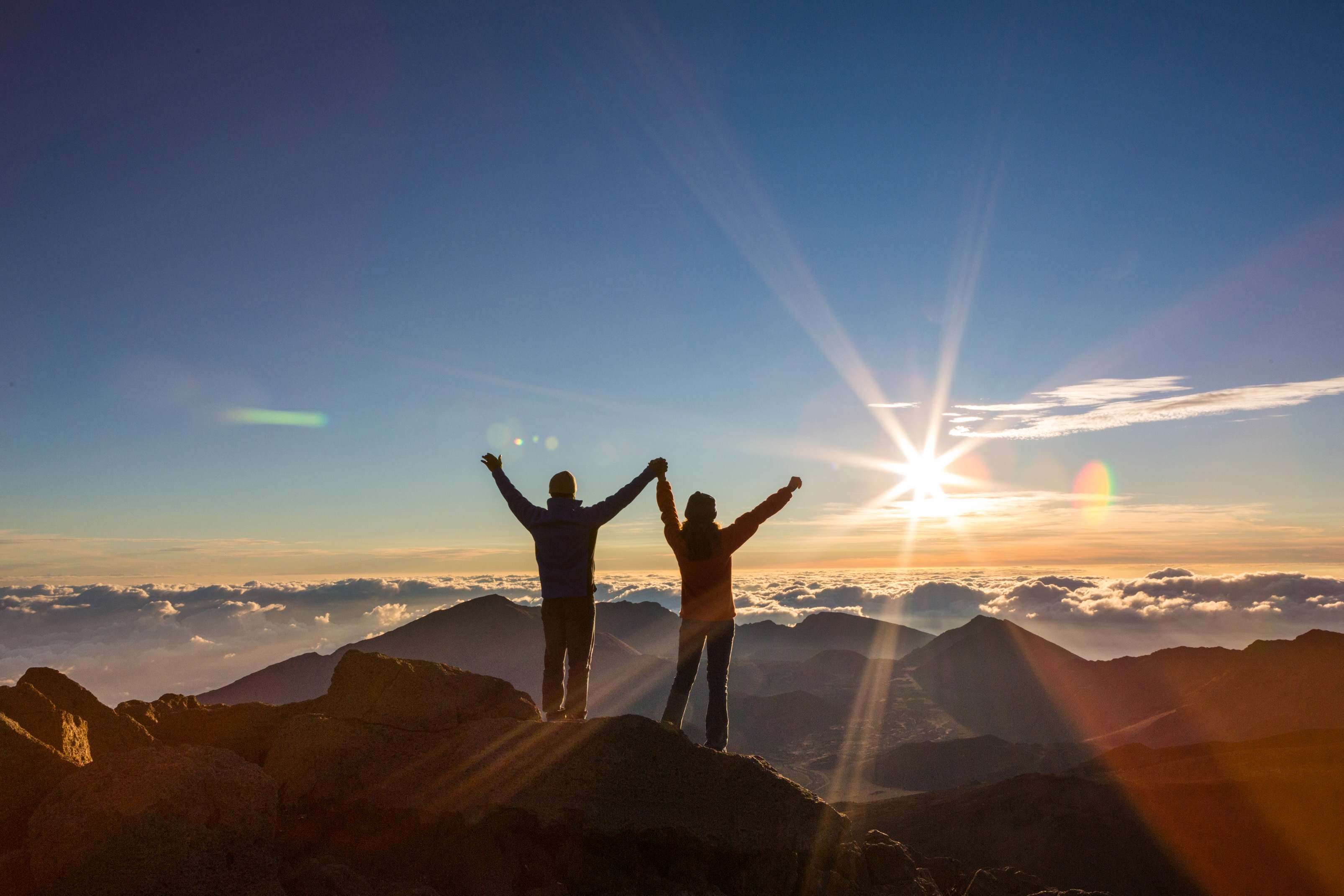 Two people standing on the summit rocks at Haleakala with arms raised as the sun clears the cloud layer