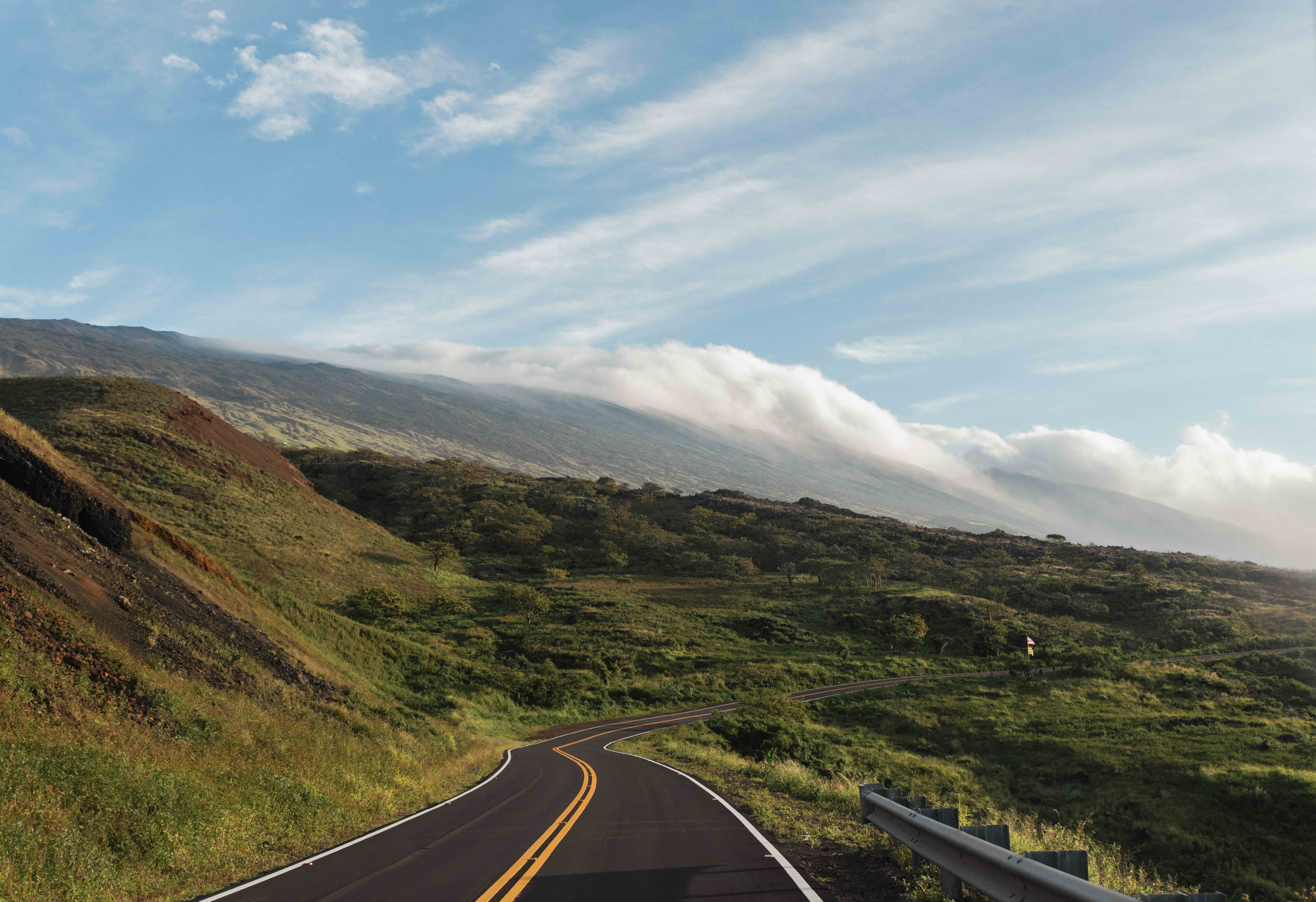 A two-lane road curving across the open upland flank of Haleakala with cloud spilling over the ridge