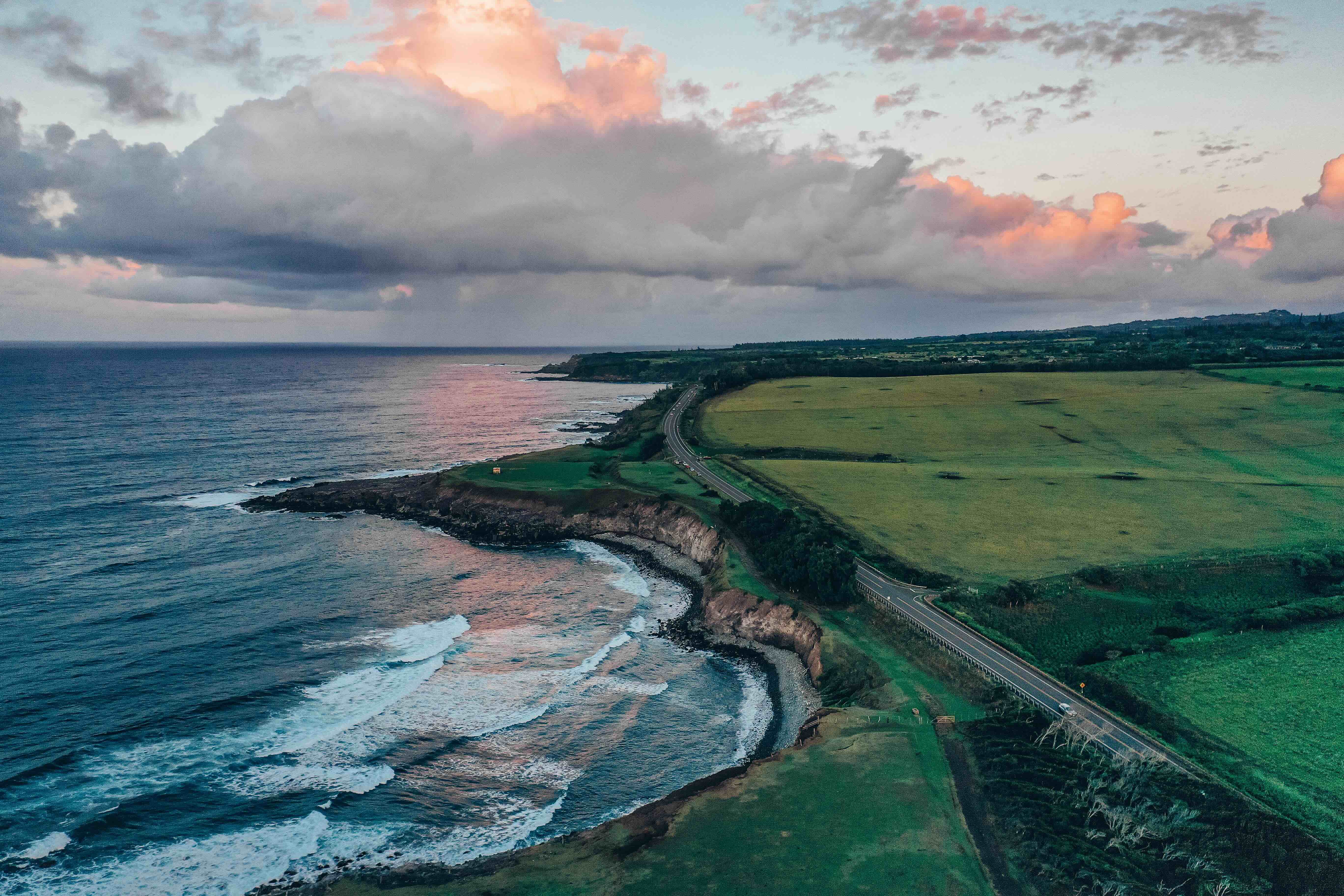 A coastal road running along green clifftop pasture above a surf-lined shoreline at sunset
