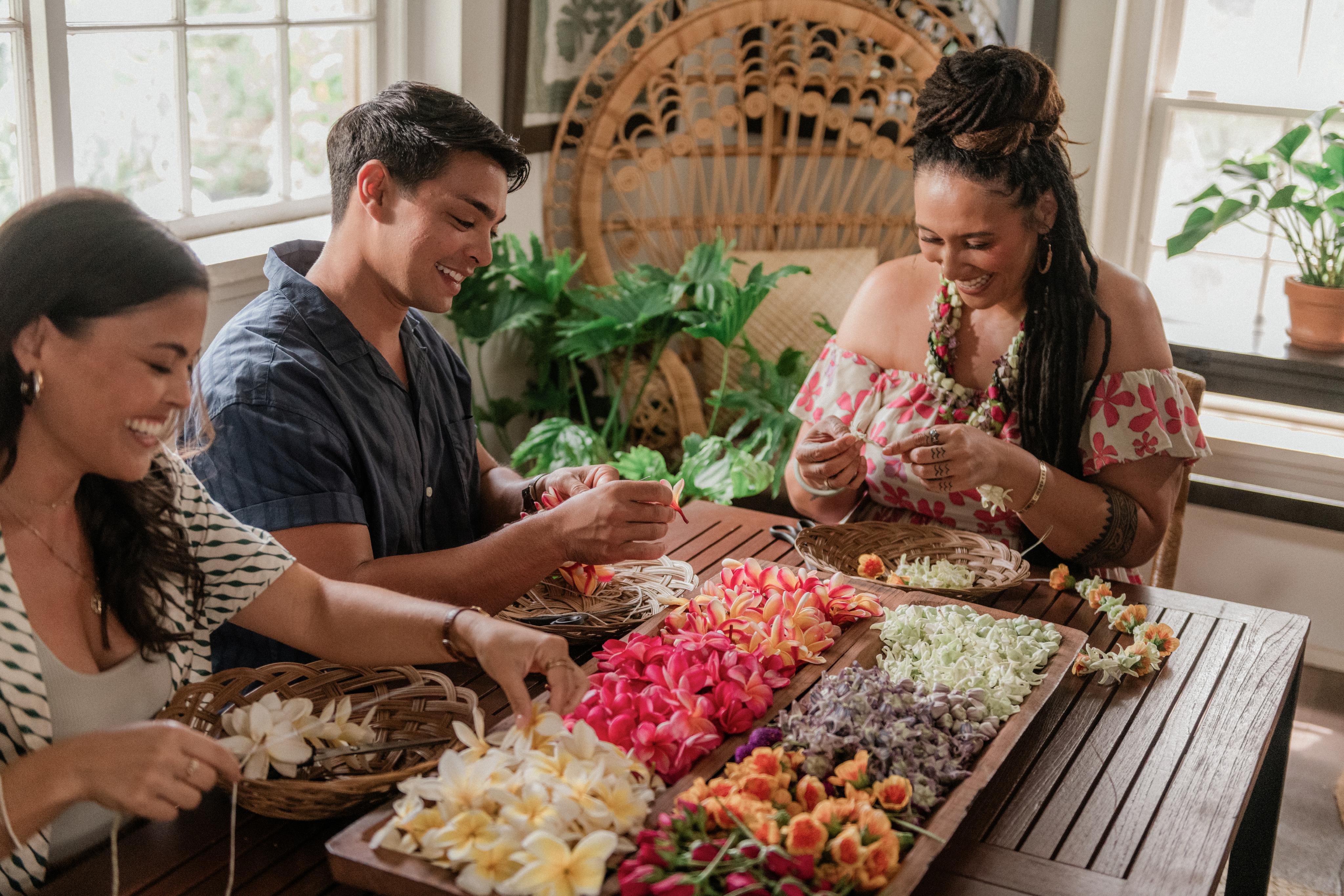 Three people making flower lei at a table covered in orchids and plumeria