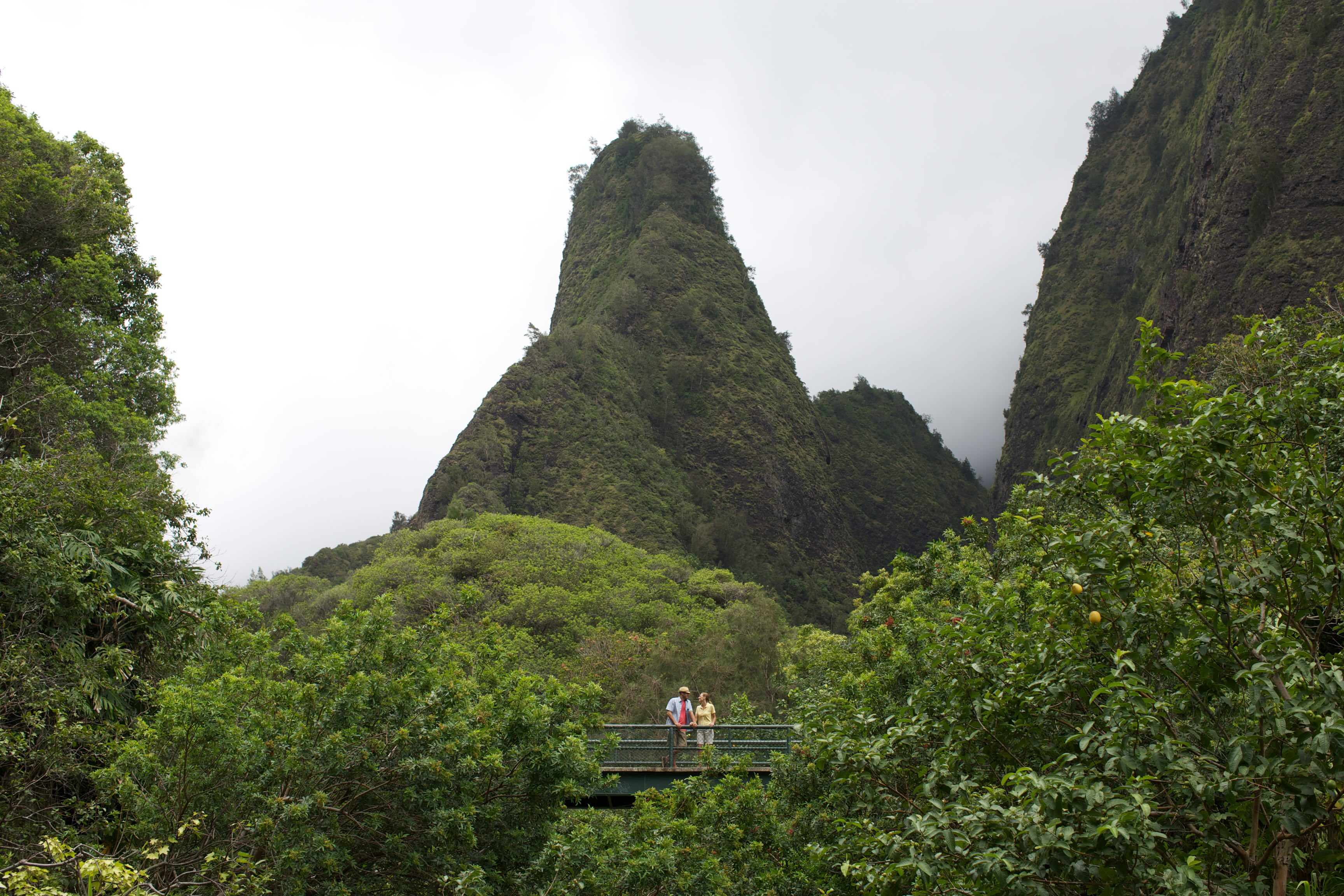 The Iao Needle rising green out of cloud in Iao Valley, with two people on the viewing bridge below