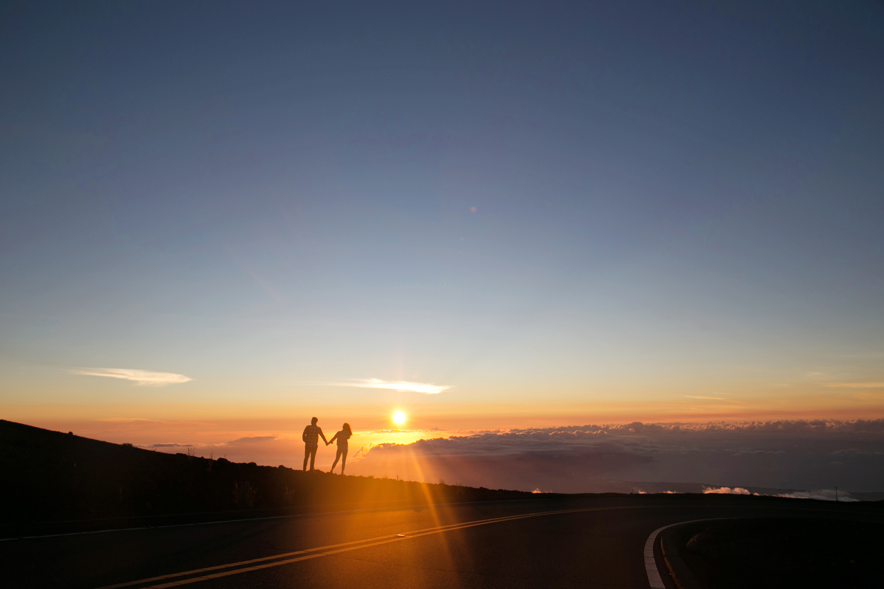 Two people standing in silhouette beside the summit road at Haleakala as the sun drops to the horizon above a low cloud bank