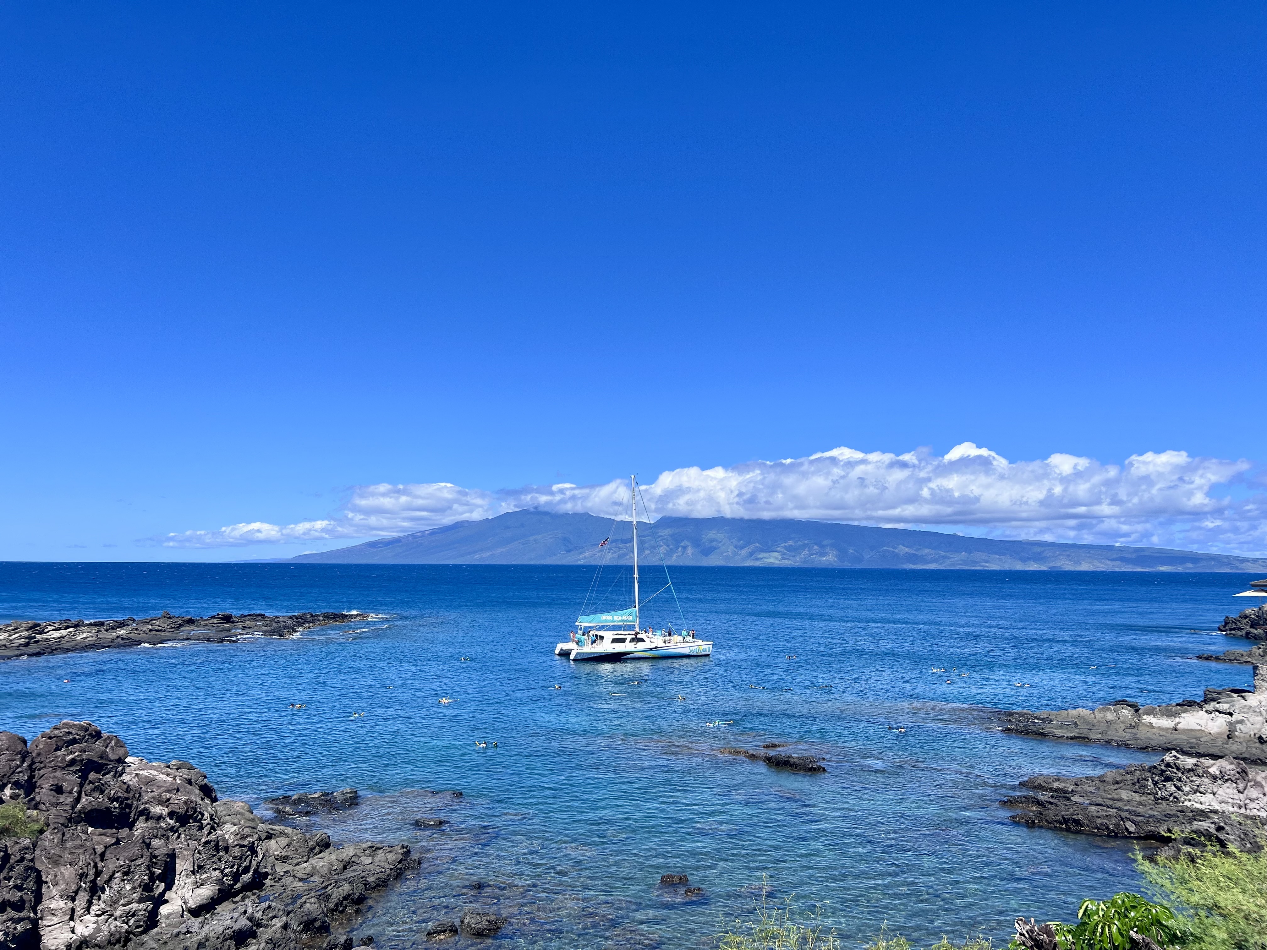 A white snorkel catamaran anchored in deep blue water beyond a lava shoreline at Kapalua, with a neighbouring island across the channel